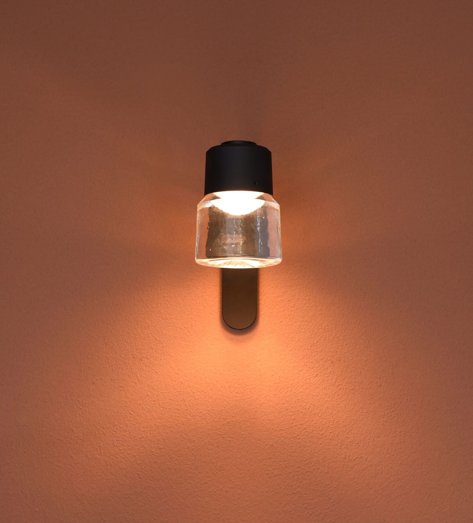 The CAST W cast glass luminaire with black aluminum body and wall bracket from a front view on a terracotta-colored wall.