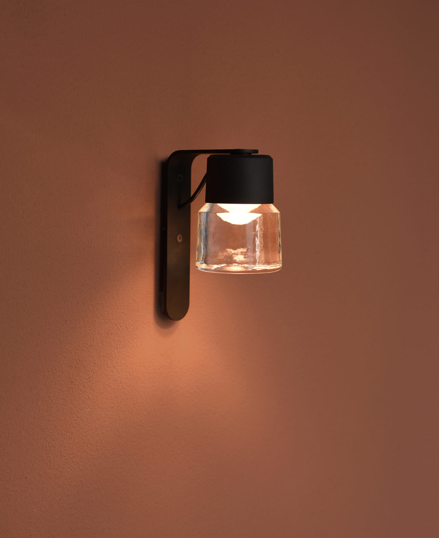 The CAST W cast glass luminaire with black aluminum body and wall bracket from a side view on a terracotta-colored wall.